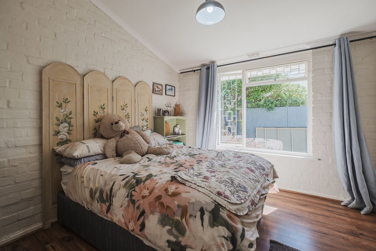 A cozy bedroom featuring painted brick walls, laminate flooring, and a large window with security bars, offering a bright and comfortable living space.