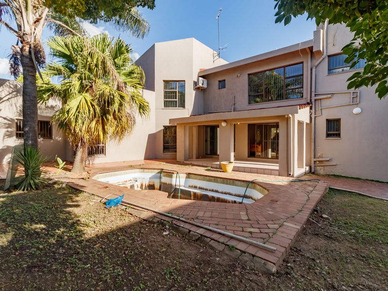 Rear view of the property featuring a covered patio and an empty swimming pool with brick paving in a garden setting.