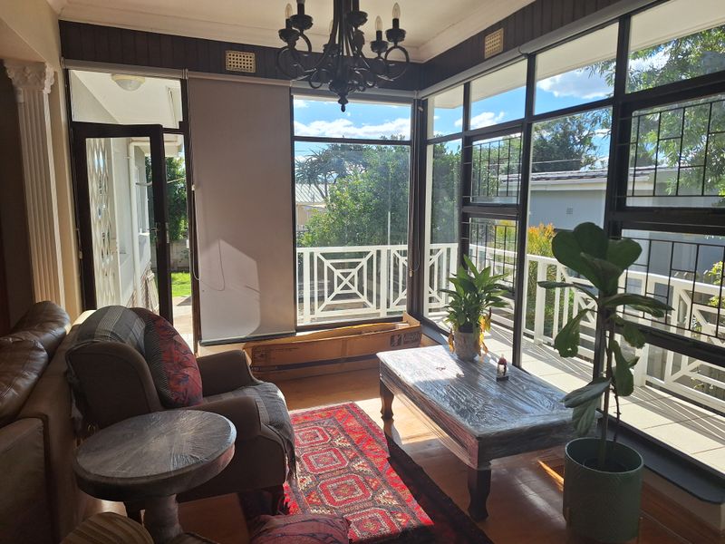 Sun-drenched living room featuring extensive glass windows, timber flooring, and a classic chandelier, offering a bright and airy atmosphere with garden views.