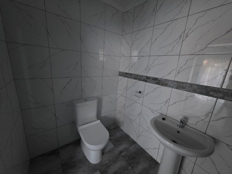 A clean, modern bathroom featuring white marble-look wall tiles, grey floor tiles, a pedestal basin, and a standard toilet.