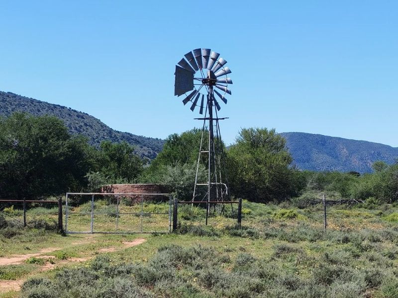 A rural farm landscape featuring a traditional metal windmill, a circular brick water reservoir, and perimeter wire fencing set against a mountain backdrop.