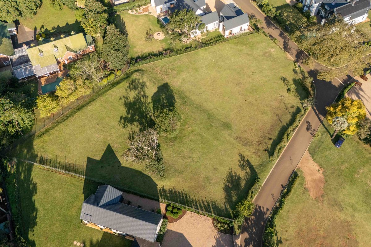 An aerial view of a vacant residential plot featuring established trees, perimeter fencing, and direct access to a paved road in a developed neighborhood.