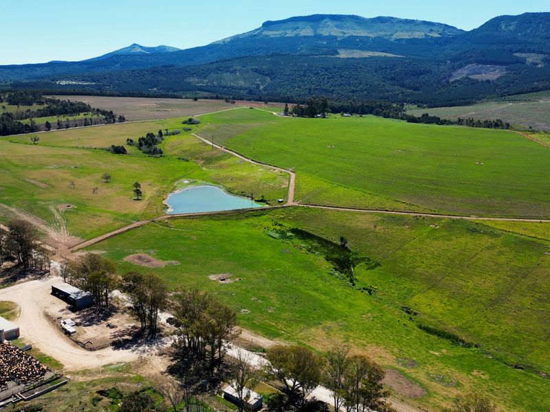 Aerial view of a large farm property featuring expansive green pastures, a central dam, livestock enclosures, and various farm infrastructure against a mountain backdrop.