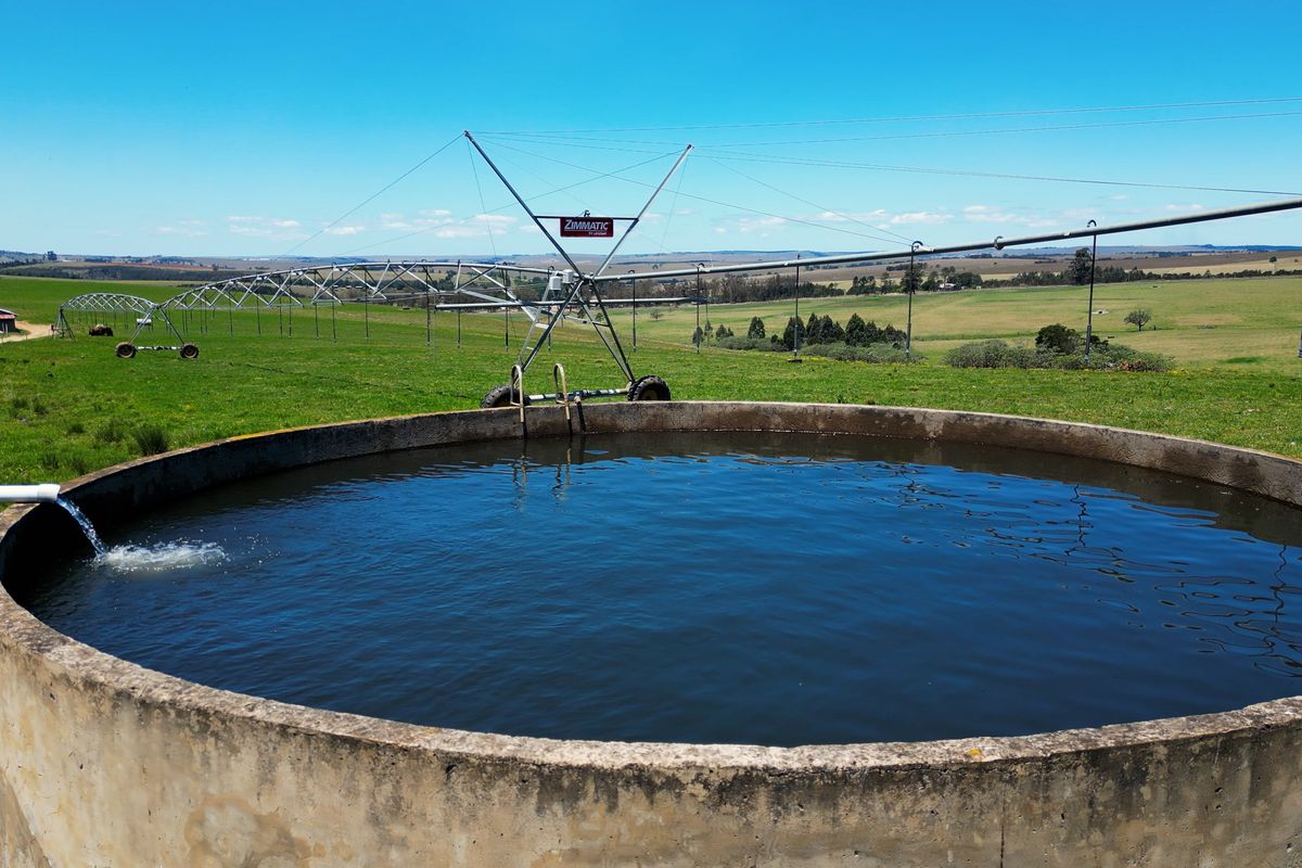 A large concrete water reservoir situated in a rural field, featuring an active center pivot irrigation system in the background.