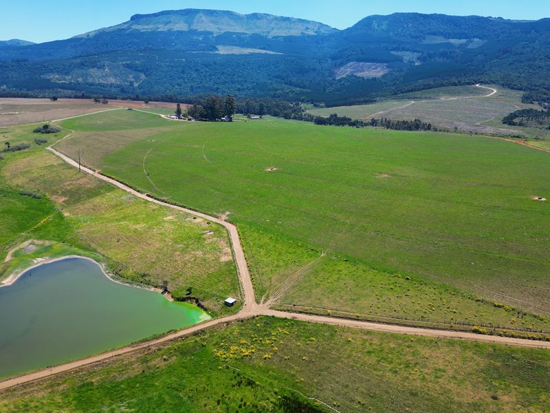 Aerial view of a large farm property featuring expansive green pastures, a dam, and gravel access roads set against a scenic mountain backdrop.
