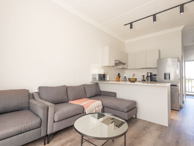 An open-plan living area featuring a modern kitchen with sleek cabinetry, track lighting, and durable laminate flooring, leading to a balcony.
