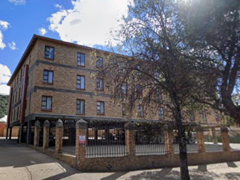 Exterior view of a multi-story face-brick townhouse complex featuring secure perimeter fencing and a paved driveway with covered parking.
