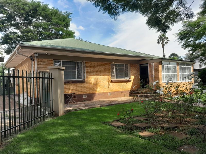 Front view of a single-story brick house featuring a metal roof, security bars on windows, and a landscaped front garden with a paved walkway.