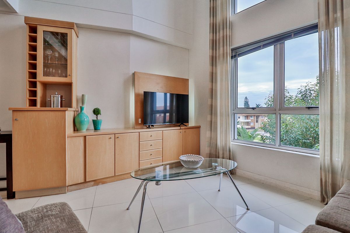 A bright living room featuring double-volume ceilings, large windows, tiled flooring, and extensive built-in wooden cabinetry.