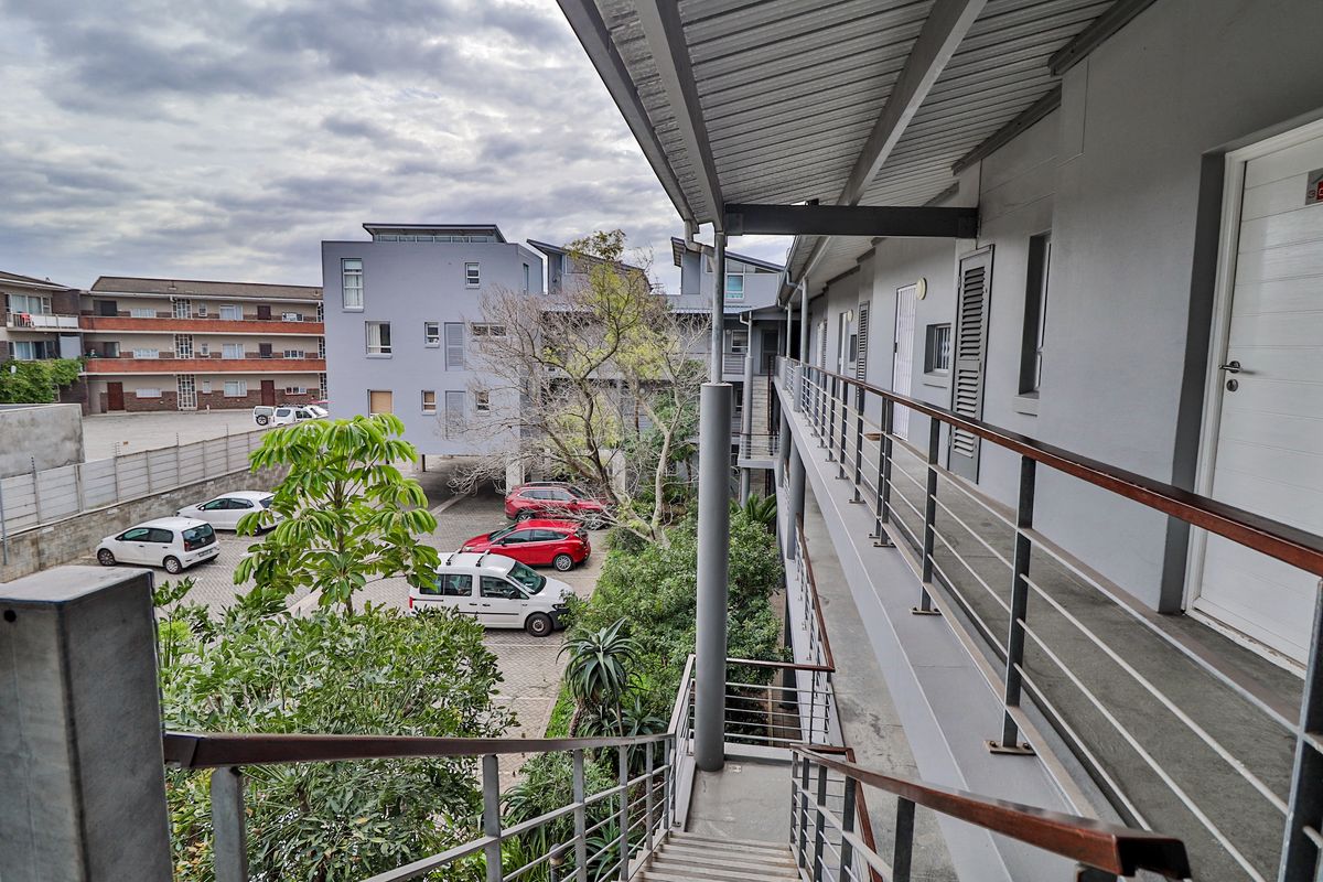An elevated exterior walkway and staircase providing access to apartment units, overlooking a paved parking area and landscaped grounds.