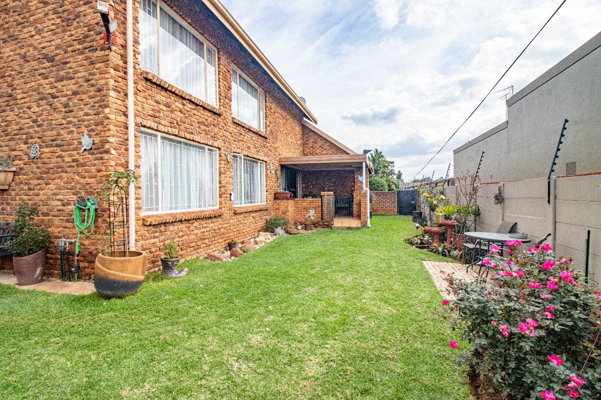 A rear view of the townhouse featuring a face brick facade, a covered patio, a well-maintained lawn, and secure boundary walls with electric fencing.