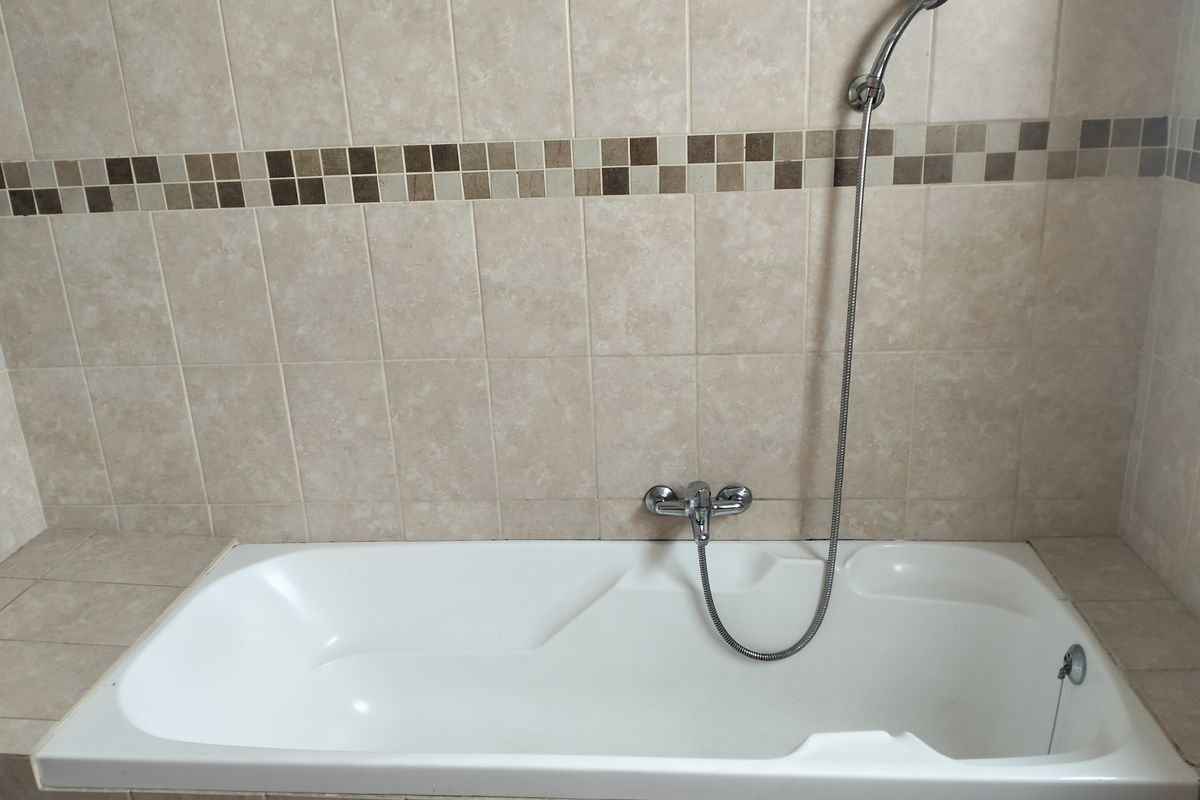 A built-in bathtub featuring neutral-toned wall tiles with a decorative mosaic border and a chrome handheld shower mixer.