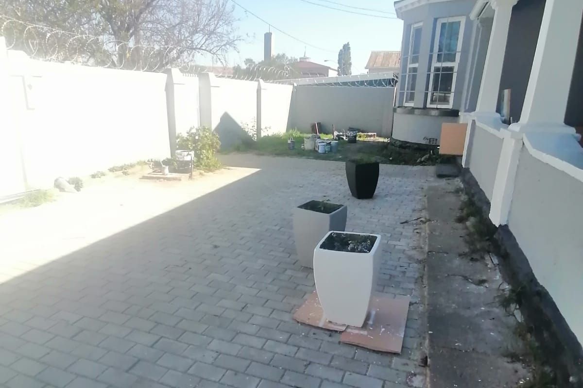 Rear exterior area featuring paved ground, perimeter wall with security fencing, and a bay window on the side of the house.