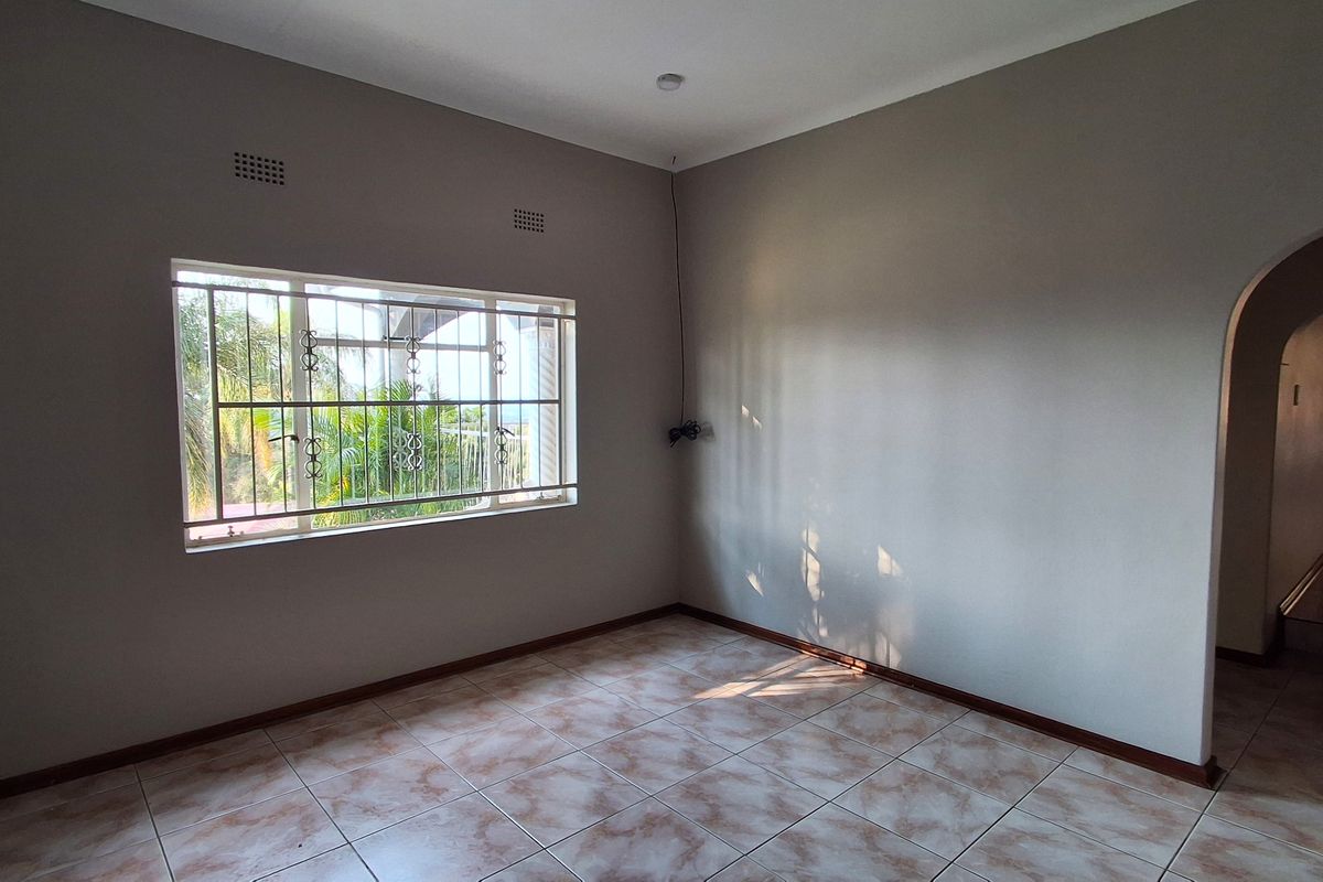 A vacant room featuring neutral wall paint, tiled flooring, and a window fitted with security bars, leading into an adjacent space via an arched doorway.