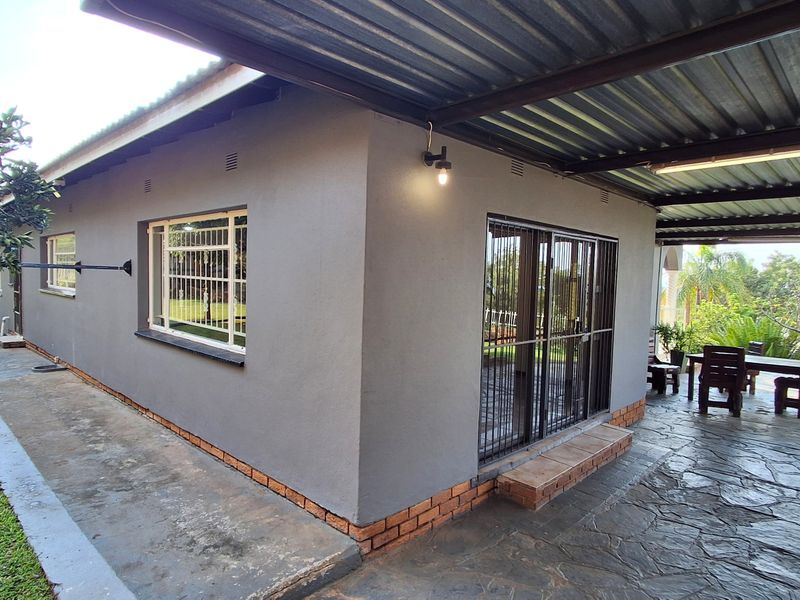A rear exterior view featuring a covered patio area with stone flooring, corrugated metal roofing, and a grey rendered facade with security bars.