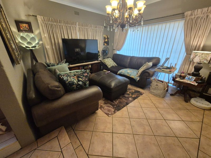 A cozy living room featuring ceramic tile flooring, a decorative chandelier, and large windows with vertical blinds, providing a comfortable and functional living space.