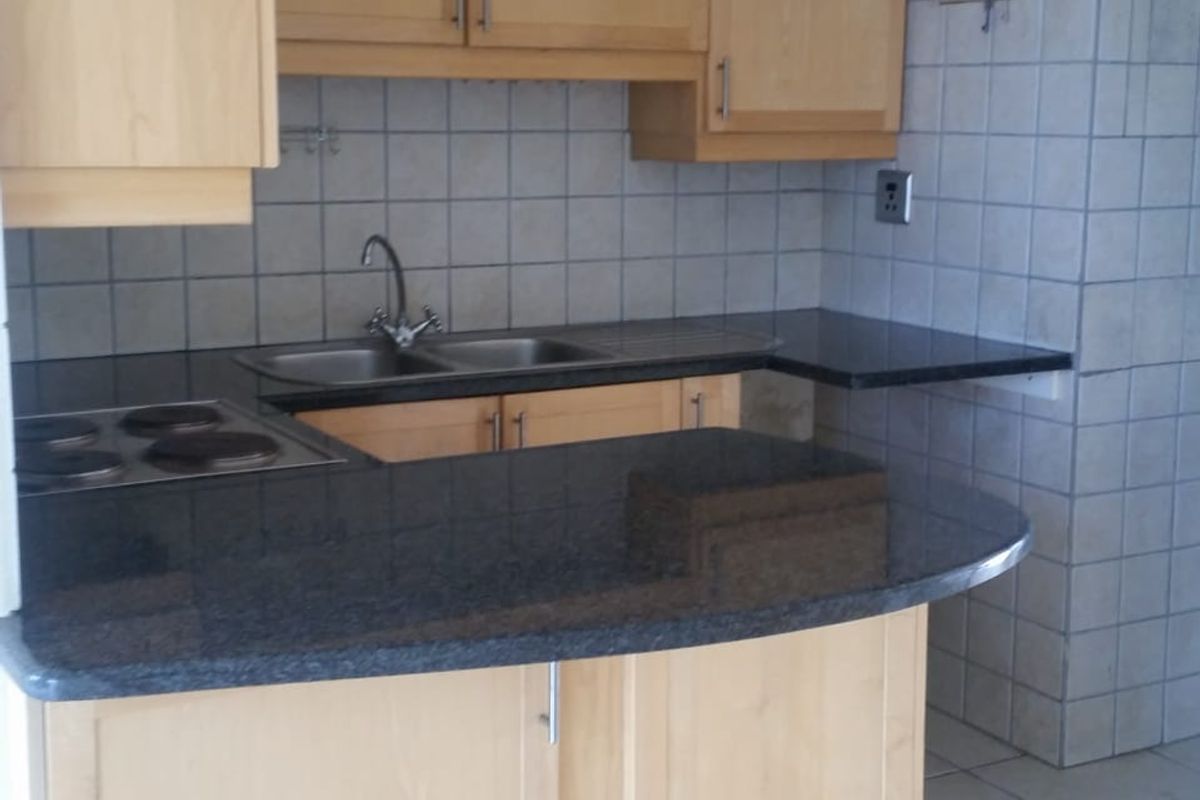A functional kitchen featuring light wood cabinetry, dark granite countertops, an integrated electric hob, and neutral ceramic tiling throughout.
