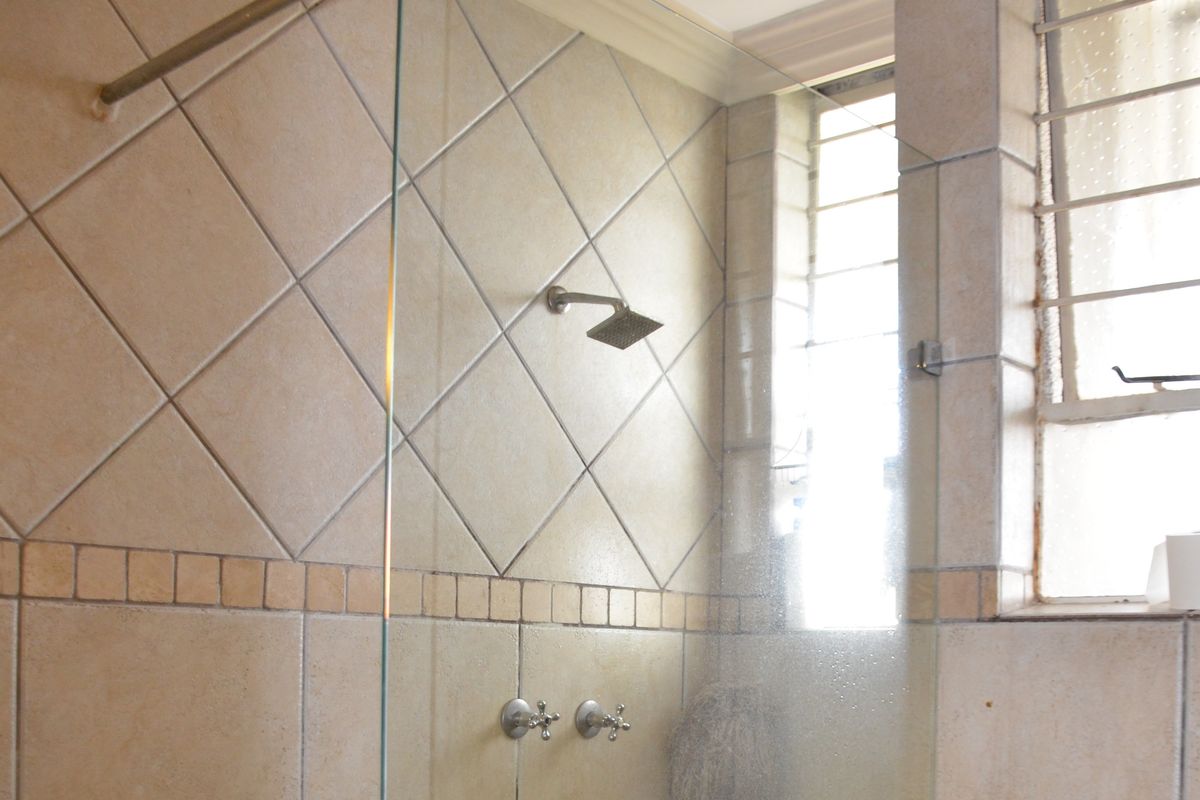 A functional bathroom shower area featuring a glass screen, wall-mounted shower head, and neutral ceramic tiling with a window for natural light.