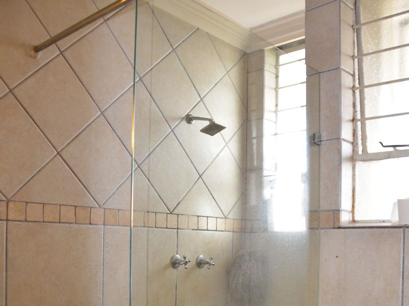 A functional bathroom shower area featuring a glass screen, wall-mounted shower head, and neutral ceramic tiling with a window for natural light.