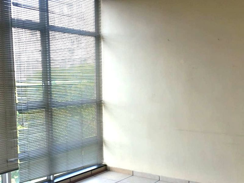 An empty living area featuring large floor-to-ceiling windows with venetian blinds and neutral ceramic floor tiles.