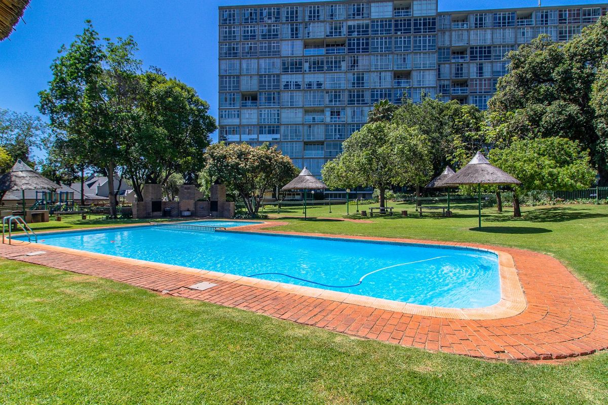 A well-maintained communal swimming pool area featuring brick coping, thatched umbrellas, and a built-in braai set within a spacious landscaped garden.