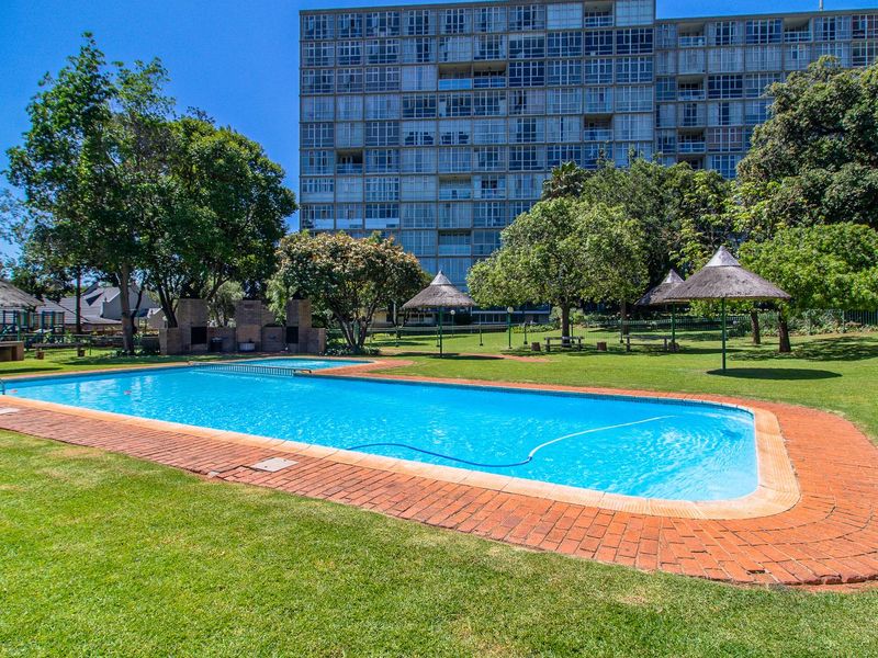 A well-maintained communal swimming pool area featuring brick coping, thatched umbrellas, and a built-in braai set within a spacious landscaped garden.