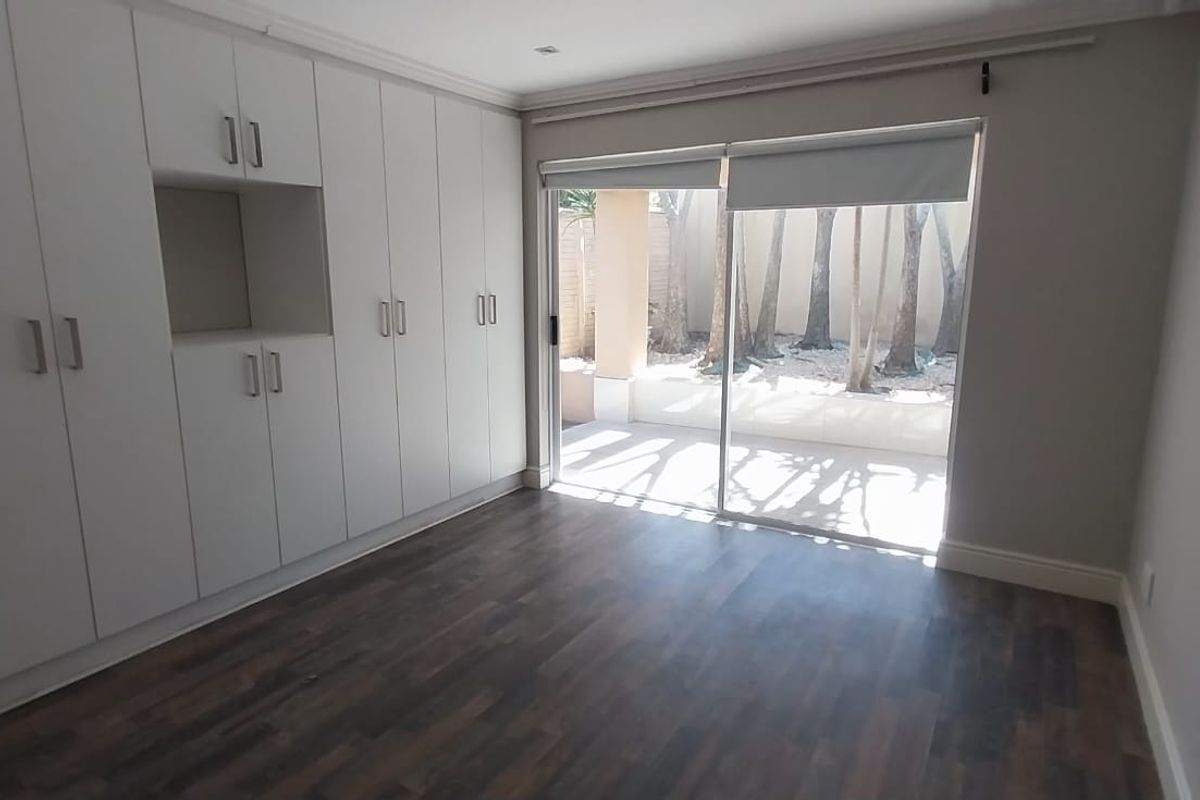 A bright, empty bedroom featuring extensive built-in cupboards, dark laminate flooring, and a sliding glass door providing access to a private outdoor area.