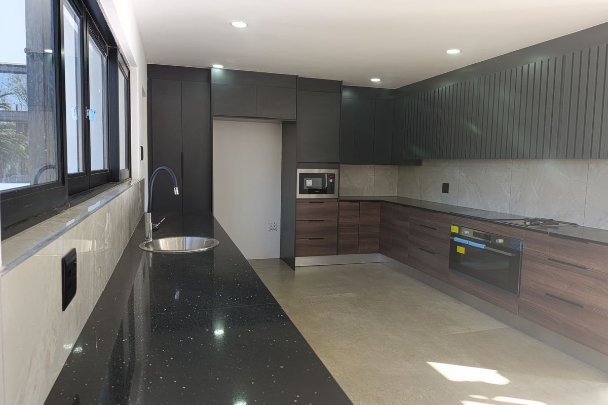 Modern kitchen featuring dark wood and matte black cabinetry, stone countertops, integrated appliances, and sleek tiled backsplash with ample natural light.