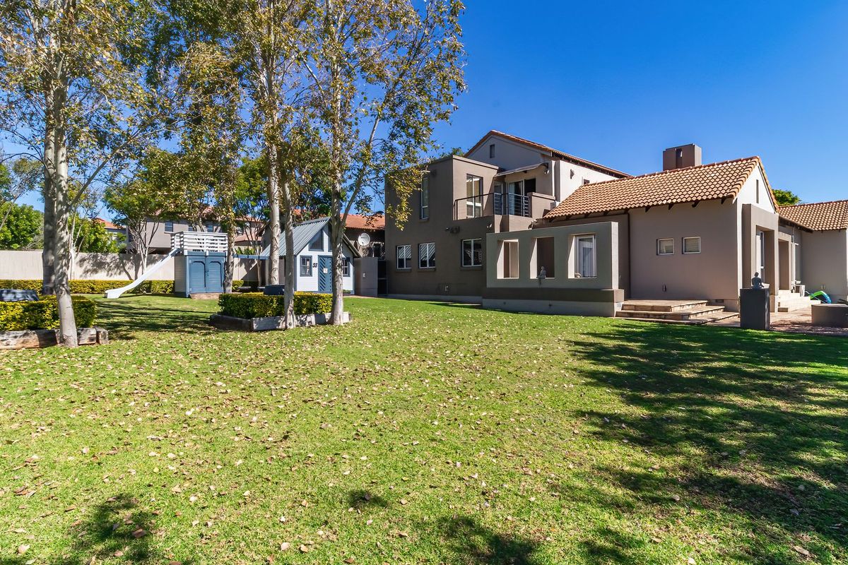 Spacious rear garden featuring a well-maintained lawn, mature trees, and a multi-level house with a balcony and paved patio area.