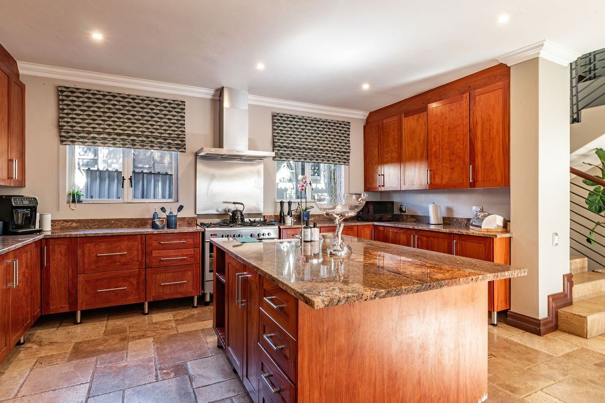 Spacious kitchen featuring warm wooden cabinetry, granite countertops, a central island, and stone tile flooring, adjacent to an open staircase.