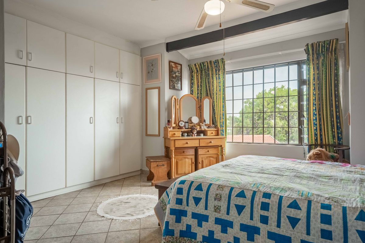 A bedroom featuring tiled flooring, extensive built-in cupboards, a ceiling fan, and a window fitted with security bars.