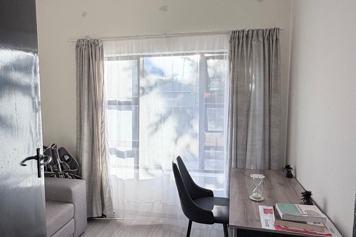 A functional bedroom or home office space featuring light-toned laminate flooring, a ceiling fan, and natural light through a window with curtain tracks.