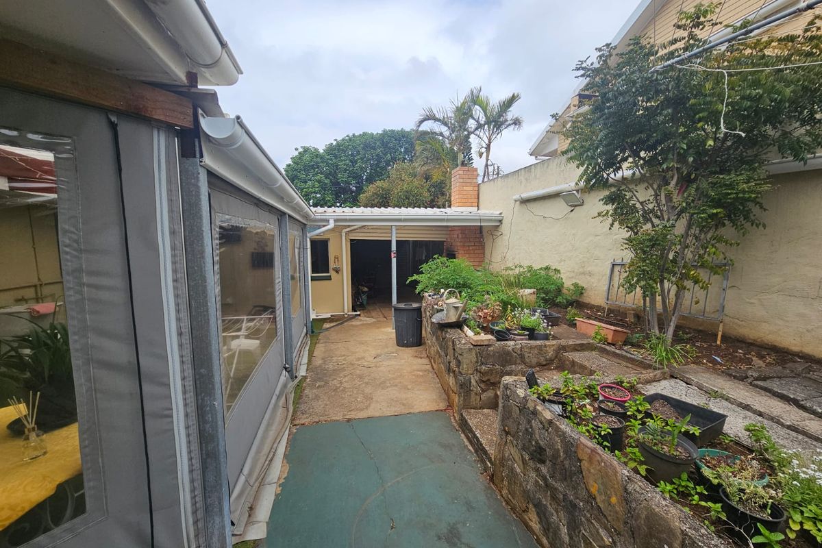 Rear exterior area featuring a covered patio, brick chimney, and tiered stone retaining walls with established garden beds.