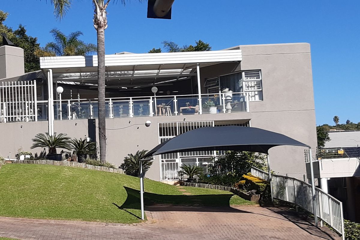 Rear view of the property featuring a paved driveway with a shade sail, a raised covered patio with glass balustrades, and landscaped garden areas.