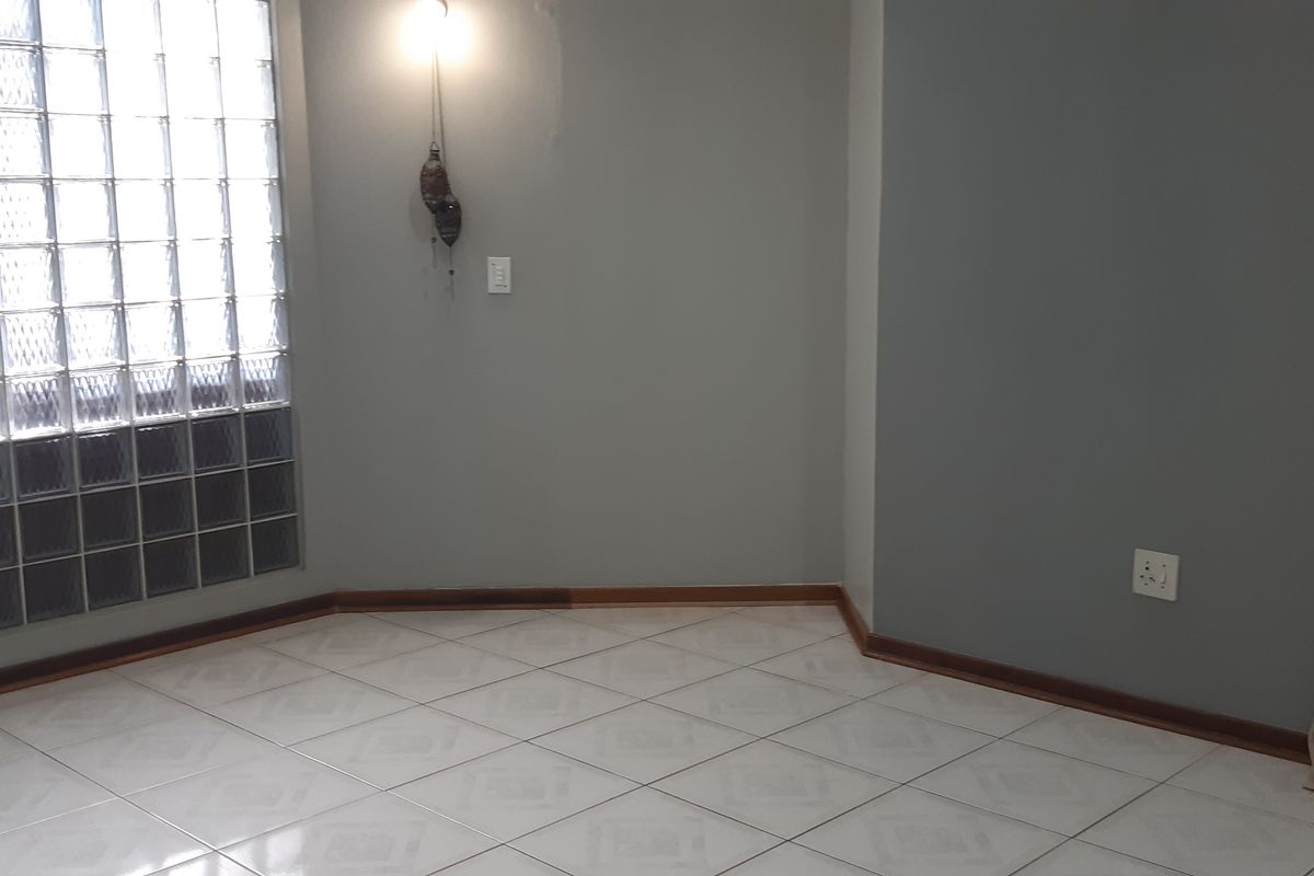 An empty living area featuring light-colored ceramic floor tiles, a distinctive glass block window, and neutral grey walls.