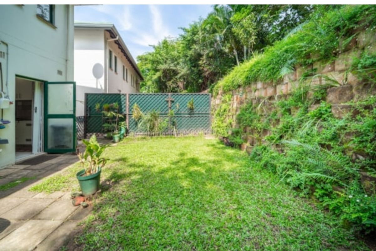A private rear garden featuring a lawn, paved patio area, and a tiered retaining wall with established greenery and perimeter lattice fencing.