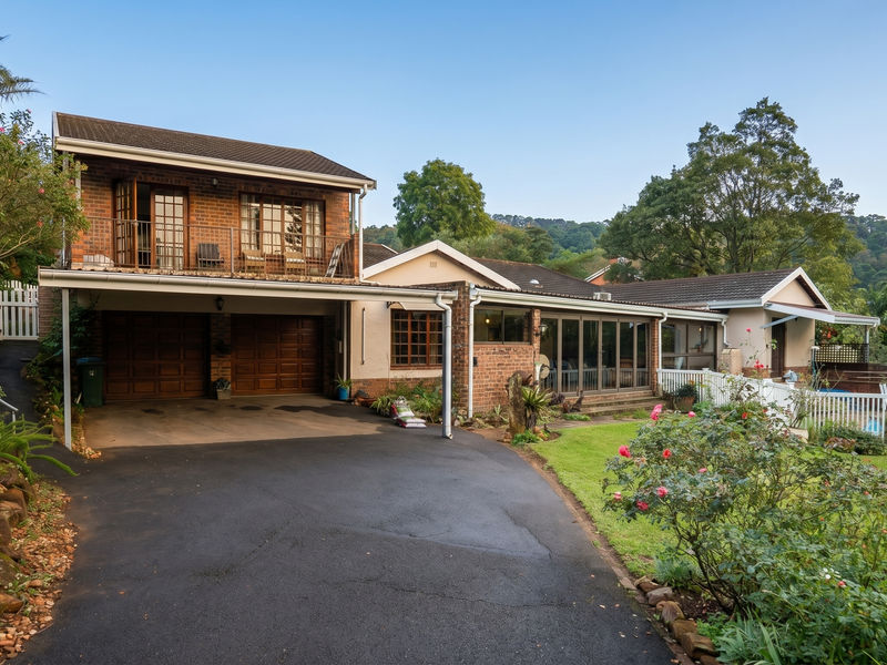 A well-maintained double-story brick home featuring a spacious asphalt driveway, double garage, upper-level balcony, and a private swimming pool area.