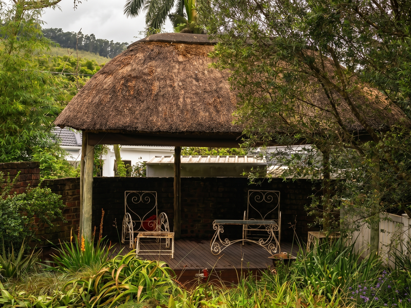A charming outdoor gazebo featuring a traditional thatched roof and timber decking, nestled within a lush, landscaped garden setting.