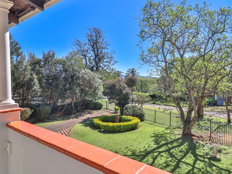 An elevated balcony overlooking a well-maintained front garden with mature trees, lawn, and metal perimeter fencing.