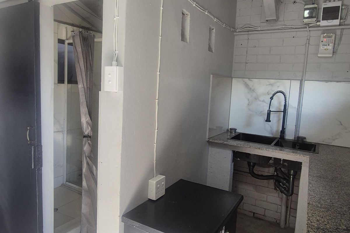 A compact kitchen area featuring granite countertops, a double basin sink, and exposed industrial-style brickwork and ceiling beams, adjacent to a small bathroom.