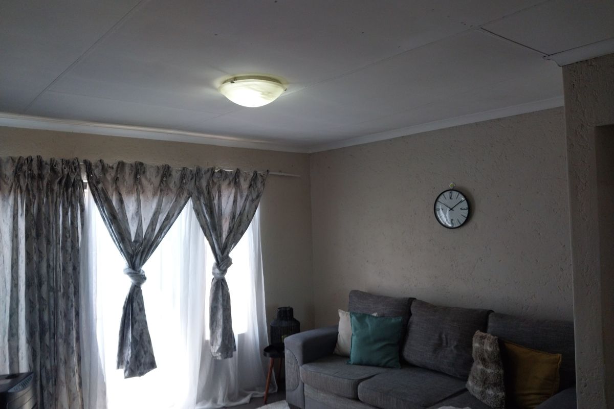 A living room featuring tiled flooring and a central ceiling light fixture, with natural light entering through large window treatments.