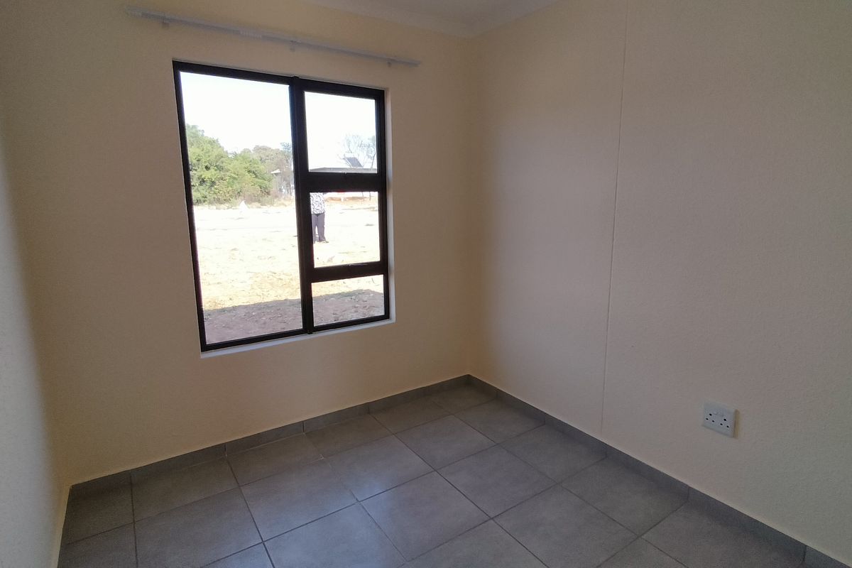 A clean, empty bedroom featuring neutral wall finishes, grey floor tiles, and an aluminium-framed window providing natural light.