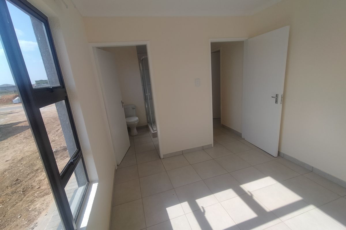 A bright, empty bedroom featuring tiled flooring, aluminium window frames, and direct access to an en-suite bathroom.