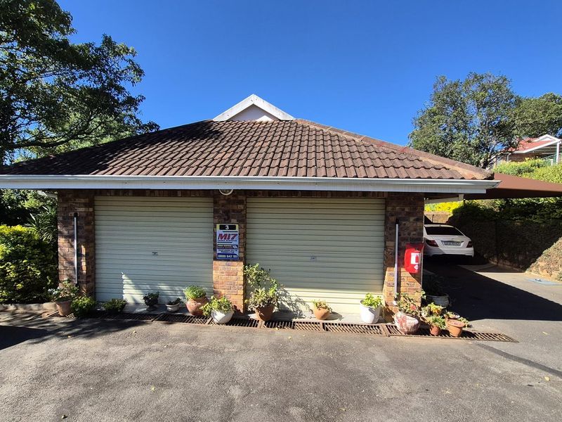 A double garage with roller doors and a tiled roof, featuring a paved driveway and established garden surroundings.
