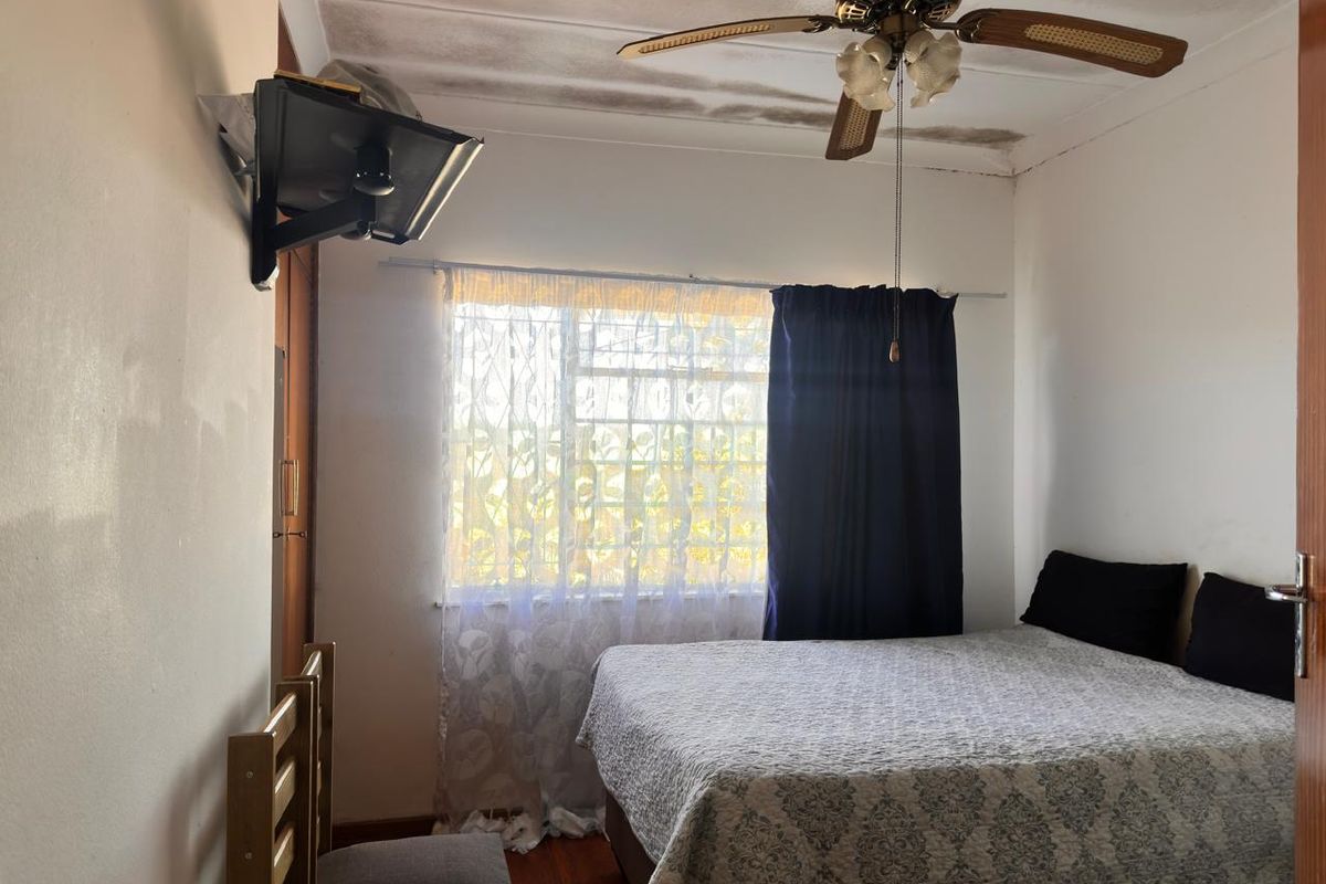 A bedroom featuring a ceiling fan, wooden flooring, and a window with security bars, currently showing signs of significant ceiling water damage.