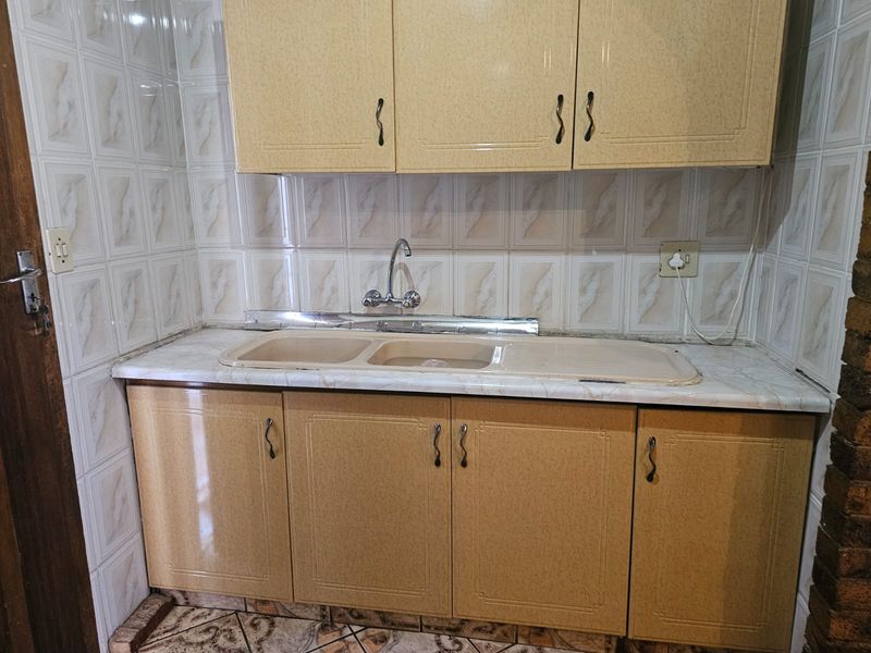 A compact kitchen area featuring dated laminate cabinetry, a double bowl sink with mixer tap, and tiled splashbacks.