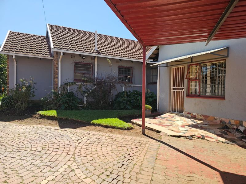 Rear exterior view featuring a brick paved driveway, metal carport, and a tiled patio area with security features on windows and doors.