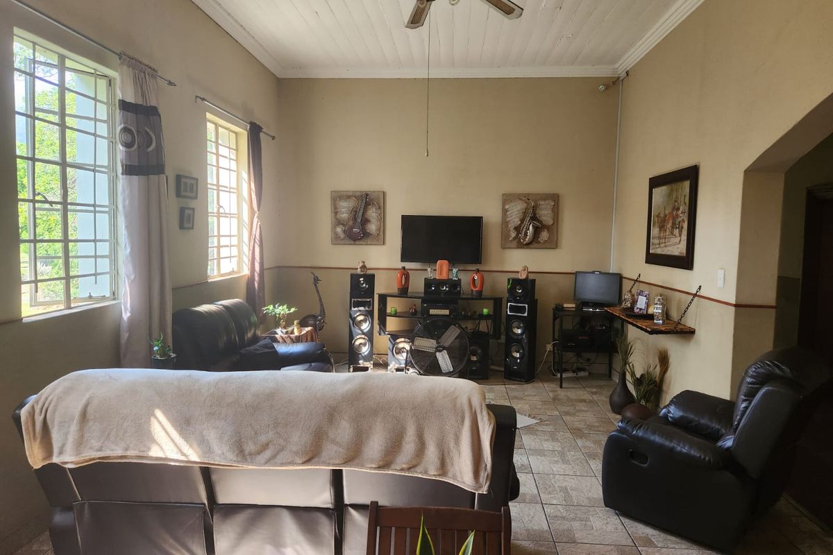 A spacious living room featuring tiled flooring, a timber panelled ceiling with a fan, and large windows with security bars.