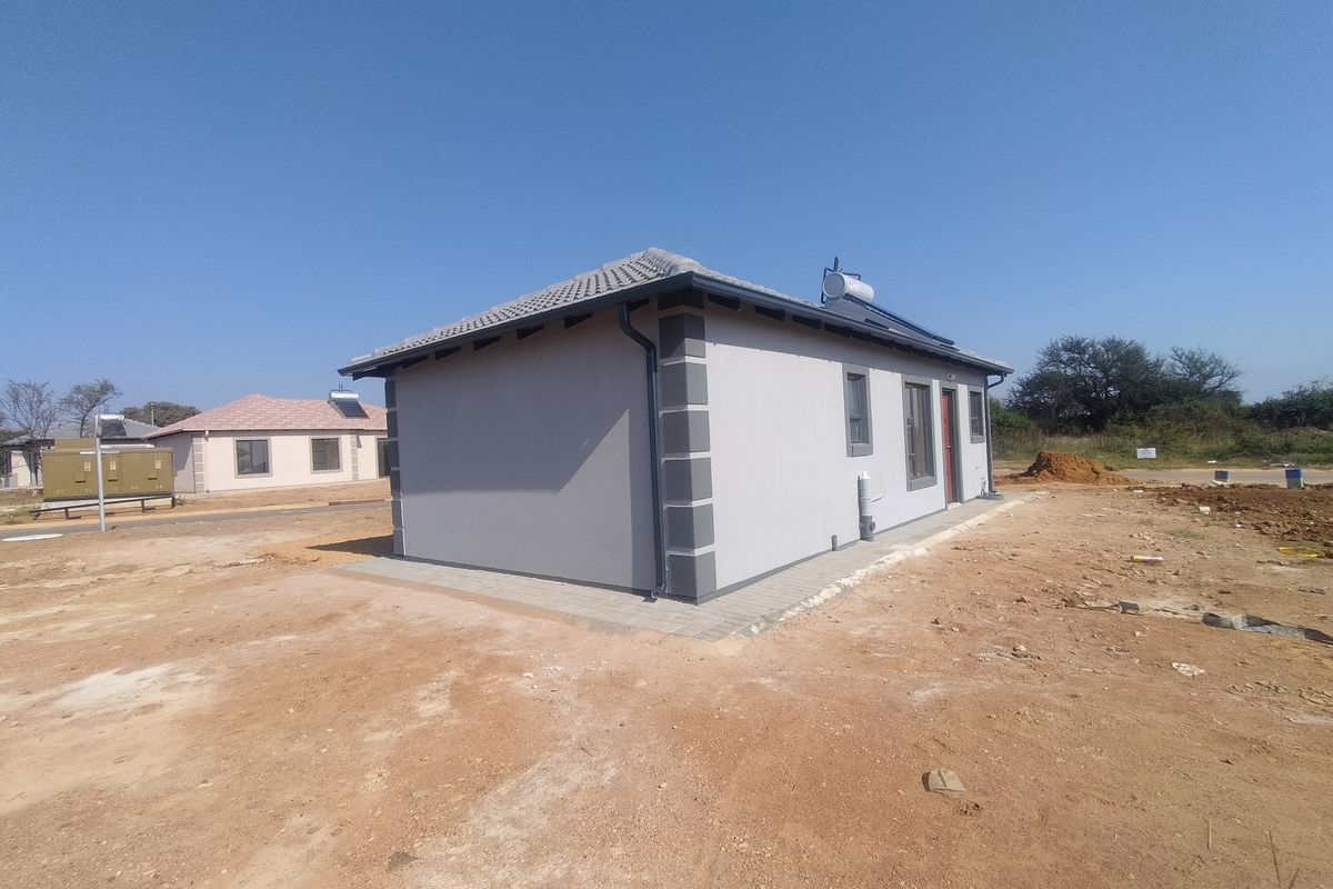 Rear view of a modern single-story house featuring a tiled roof, solar water heating system, and paved perimeter on an undeveloped plot.