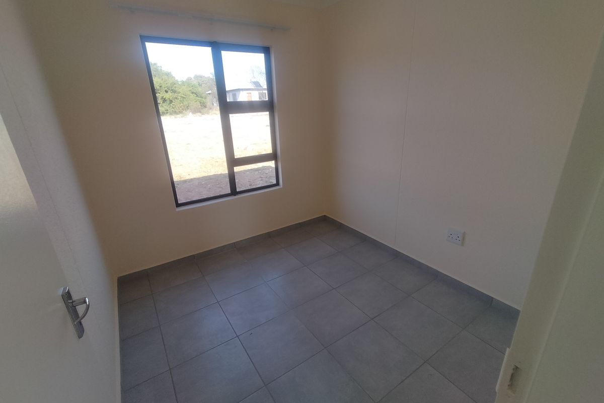 A clean, empty bedroom featuring neutral wall paint, grey floor tiles, and a large aluminium-framed window providing natural light.